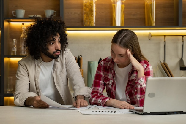 A worried-looking couple sitting at a table with financial documents, calculators, and a laptop displaying a graph showing rising interest rates. The setting is a dimly lit home office.